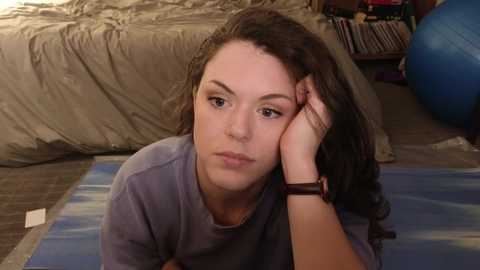 Media: A video of a young woman with curly hair, wearing a purple shirt, lying on a blue mat, looking contemplative in a messy bedroom with a bed and a blue exercise ball in the background.