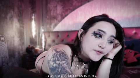 Media: Video of a pale-skinned woman with dark hair and black eyeliner, wearing a black choker, leaning on a bed with a tufted headboard in a dimly lit, gothic-style room.