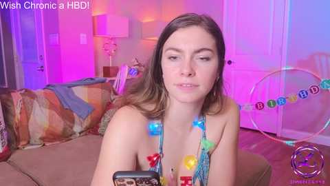 Media: Video of a young woman with light skin, brown hair, and a neutral expression, wearing a colorful beaded necklace and a low-cut, revealing top, sitting on a brown couch in a brightly lit, colorful room with a birthday-themed backdrop.