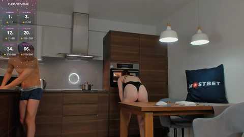 Media: Video of a woman in black lingerie bent over a kitchen counter, revealing her buttocks, while a shirtless man stands nearby in a modern kitchen.