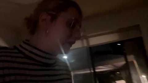 Media: Video of a woman with shoulder-length brown hair, wearing a black and white striped turtleneck, leaning against a window, looking outdoors. Dimly lit room with a soft glow from the streetlight outside.