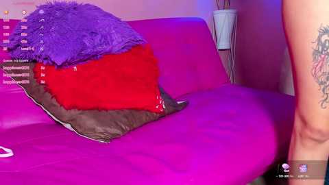 Media: Video of a bright pink bed with a fluffy purple pillow and a red pillow, featuring a person's tattooed thigh.