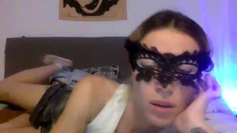 Media: Video of a woman with light skin, brown hair, and wearing a black lace masquerade mask, lying on a bed with a grey headboard, wearing a white tank top, and grey shorts.