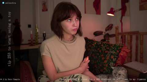 Media: Video of a young woman with shoulder-length brown hair, wearing a beige T-shirt, sitting on a bed with floral pillows in a cozy bedroom.