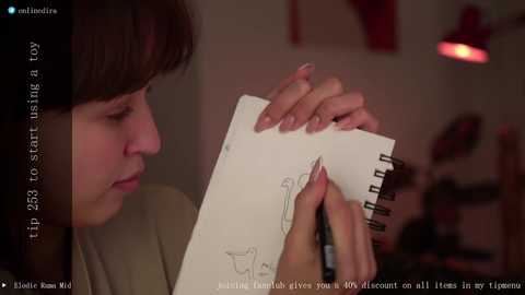 Media: A video of a woman with short brown hair, holding a sketchbook, illustrating a nude female figure. The background is blurred, showing a dimly lit room with red lighting. The woman is dressed in a beige top.