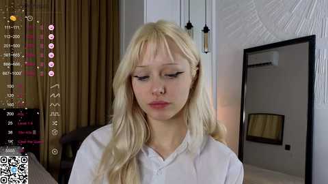 Media: Video of a blonde woman with fair skin, wearing a white shirt, standing indoors. The background features a modern room with curtains, a mirror, and a bed.