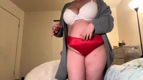 Media: Video of a light-skinned woman in a red bra and high-waisted red panties, with a gray cardigan open, standing in a beige-tiled bathroom.