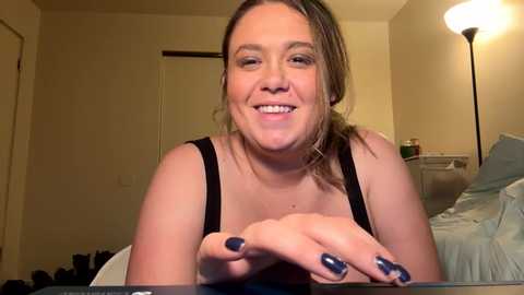 Media: Video of a smiling, light-skinned woman with medium-length brown hair in a simple black tank top, resting her hands on a table, revealing blue-painted nails. The background shows a dimly lit bedroom with a bed and lamp.