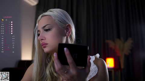 Media: A video of a blonde woman with long hair, wearing a white lace top, looking at a smartphone in a dimly lit room with dark curtains and a blurred background.