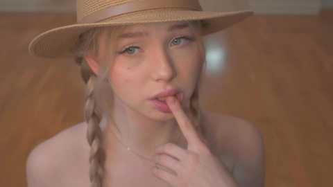 Media: Video of a fair-skinned young woman with blonde braids, wearing a straw hat, touching her lips suggestively. Background is a wooden floor, blurred, creating a soft focus effect.