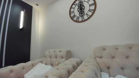 Media: Video of a minimalist, modern bedroom with beige tufted headboard, white bedding, black wall-mounted clock, and black door with vertical stripes.