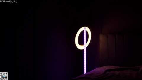 Media: Video of a dimly lit bedroom with a glowing neon ring lamp on a stand, casting a soft purple hue. The bed, visible in the lower left corner, is draped in dark sheets. The background is dark and shadowy, creating a moody, intimate atmosphere.