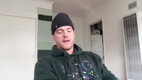 Media: Video of a young, fair-skinned man with short brown hair and a beard, wearing a dark green hoodie with a colorful graphic, and a black beanie, standing indoors in a bright room with white walls and a window.