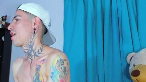 Media: Video of a shirtless, light-skinned man with a white baseball cap, tattoos, and earrings, looking to the left, against a blue curtain backdrop.