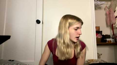 Media: Video of a young Caucasian woman with long blonde hair, wearing a maroon top, sitting in a cluttered bedroom with white walls, a closet door, and hanging clothes.