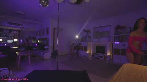 Media: Video of a dimly lit, modern living room with a stripper pole, purple lighting, and a woman in a pink off-shoulder top dancing provocatively.