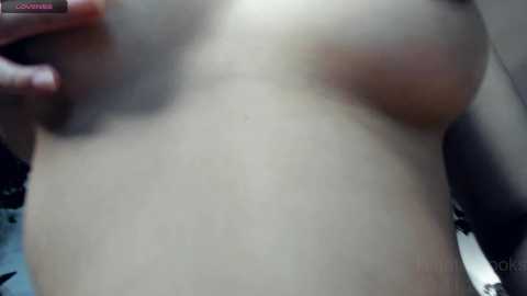 Media: A close-up video of a topless woman with fair skin, small breasts, and a hand covering one breast. The background is blurred, possibly indoors.