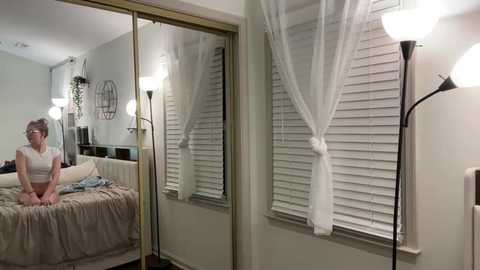 Media: Video of a woman with light skin and short brown hair, wearing a white top, sitting on a bed with beige sheets. The room has white blinds, a mirror, and a lamp.