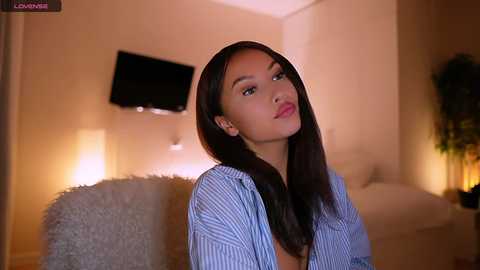 Media: Video of a young East Asian woman with long black hair, wearing a blue striped shirt, sitting in a cozy, dimly lit bedroom with a TV and plants in the background.