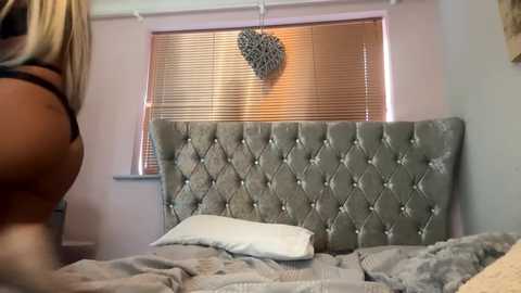 Media: Video of a cozy bedroom with a plush, tufted grey headboard, a heart-shaped wall decoration, and a blonde woman partially visible, wearing a black bra.