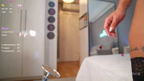 Media: Video of a woman's bare arm holding a cigarette, standing in a modern, minimalistic bedroom with white walls, wooden floors, and a bed with white sheets.