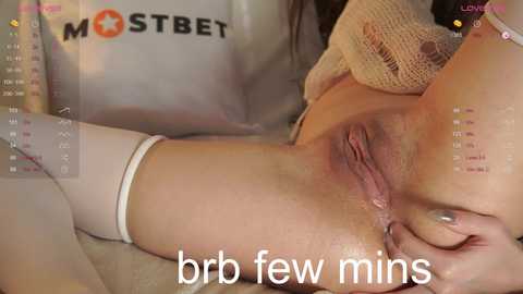 Media: Video of a light-skinned woman lying on her back, revealing her shaved vulva. The background features a white cloth with a \"MOSTBET\" logo. Text reads \"brr few mins.\