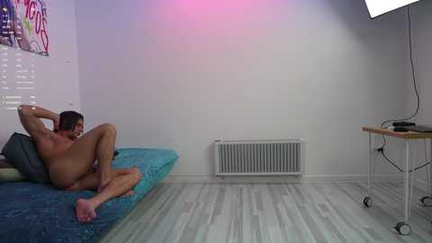 Media: Video of a nude, muscular man with short dark hair, lying on a blue bed in a minimalist room with white walls, light wooden floor, radiator, and a table with a monitor.