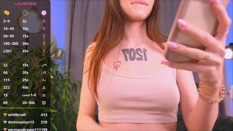 Media: Video of a fair-skinned woman with long brown hair, wearing a tight, light-pink crop top with \"YOSI\" in bold letters. She's taking a selfie with a phone in her right hand.