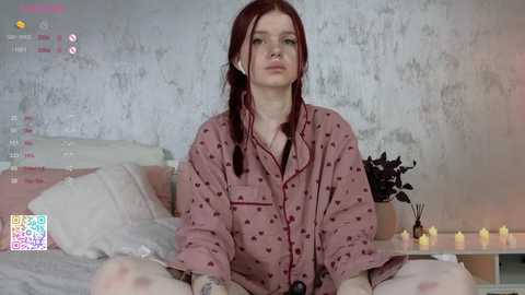 Media: Video of a pale-skinned woman with red hair in a ponytail, wearing a pink button-up shirt, sitting cross-legged on a bed with white sheets, surrounded by candles.