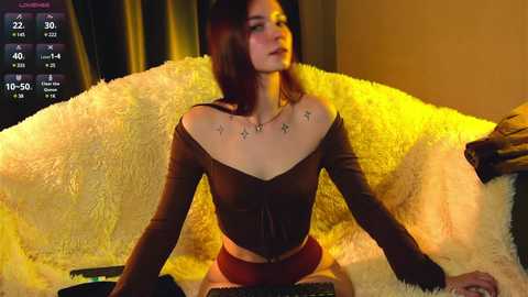 Media: Video of a fair-skinned, slim woman with long brown hair, wearing a brown off-shoulder top and red shorts, sitting on a yellow fur blanket, looking at a TV screen displaying a virtual reality game.