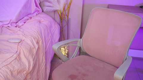 Media: A video of a beige recliner chair with a pink cushion and matching fabric, next to a bed with a pink quilt and white pillow, under a purple light.