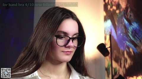 Media: Video of a young woman with long brown hair, wearing black-rimmed glasses and a white top, gazing downward in a dimly lit room with a colorful abstract painting on the right.