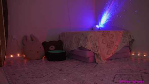 Media: A dimly lit video of a child's bedroom with a bed covered in a lavender quilt, featuring a plush bear and a stuffed animal. A blue and purple laser light projects a starry effect on the wall.