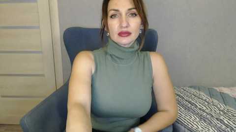 Media: Video of a fair-skinned, middle-aged woman with brown hair, wearing a sleeveless green turtleneck. She sits in a blue armchair against a gray wall, with a striped blanket on the right.