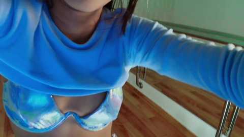 Media: A video of a light-skinned woman in a blue tie-dye crop top and matching bikini bottoms, leaning against a wooden railing in a brightly lit room with hardwood floors.