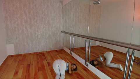 Media: Video of a ballet studio with wooden floors, mirrored walls, and a barre. A woman in white tights and a tank top is stretching on the floor, her reflection visible in the mirror.