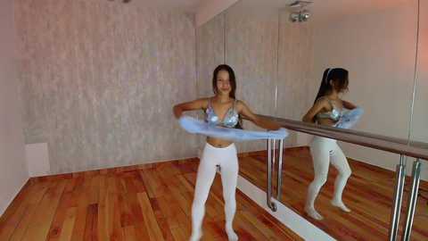 Media: Video of a young woman with medium brown skin and long black hair in a white leotard and white leggings practicing ballet on a wooden floor in a mirrored studio.