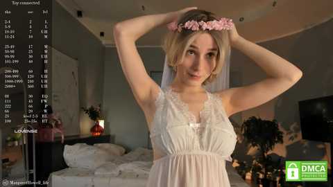 Media: Video of a young, slender, fair-skinned woman with blonde hair adorned with a pink flower crown, wearing a white lace camisole, standing in a dimly lit bedroom.