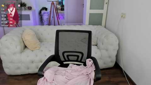 Media: Video of a white, tufted sofa with a beige pillow, a black mesh office chair with a pink blanket, and a potted plant in a modern living room.