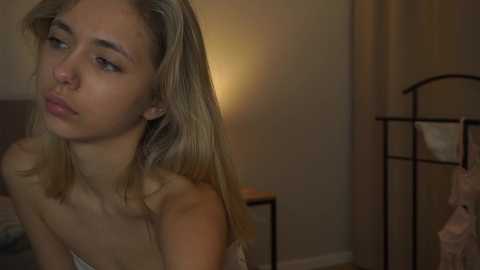 Media: A video of a young, light-skinned blonde woman with straight hair, wearing a strapless top, looking thoughtful in a dimly-lit bedroom with beige walls, a metal shelf, and a soft light.