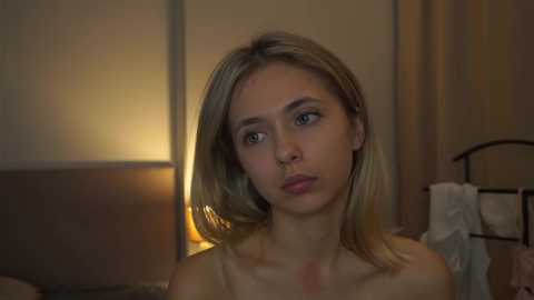 Media: A video of a blonde Caucasian woman with a light complexion and a slightly puffy face, possibly from crying, wearing a light-colored top. The background is a dimly lit bedroom with beige curtains and a warm, ambient light.