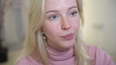 Video of a blonde woman with fair skin and blue eyes, wearing a pink turtleneck, with a blurred background.