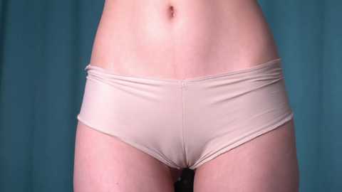 Media: Video of a woman's waist and lower abdomen, wearing beige panties against a teal background. The image focuses on the smooth skin and minimalistic garment, emphasizing the contours of her body.