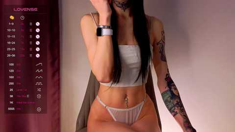 Media: Video of a slender woman with long black hair, wearing a white crop top and thong, sitting on a chair. She has tattoos on her arm and wrist. Background includes a maroon curtain and a \"LoveSense\" app interface.