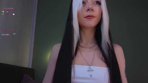 Media: Video of a young woman with long, straight black hair featuring white streaks, wearing a strapless white top and silver necklace with a cross pendant. Background is dark green.