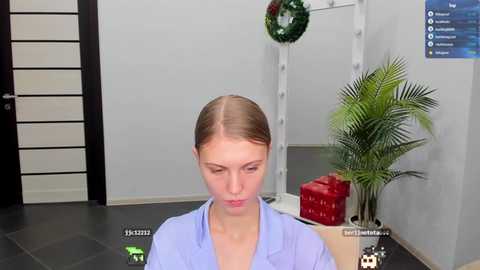 Media: A video of a young woman with fair skin and blonde hair in a ponytail, wearing a light blue shirt, standing in a minimalist room with a potted plant, a red gift box, and a Christmas wreath.