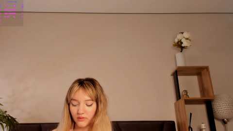 Media: Video of a young woman with long blonde hair, wearing a beige top, sitting indoors. Background includes beige walls, a white vase with white flowers, and a wooden shelf with a potted plant.