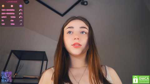 Video of a young Caucasian woman with long, straight brown hair, wearing a sleeveless top, in a modern room with a black bookshelf, green plant, and digital display.