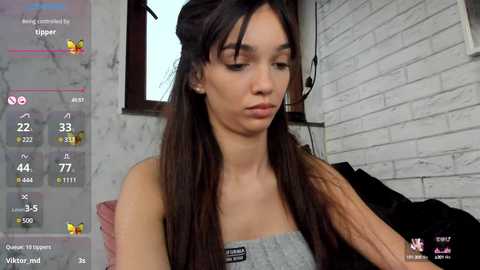 Media: Video of a young woman with long dark hair, wearing a gray off-shoulder top, sitting in a modern room with white brick walls and a pink couch.