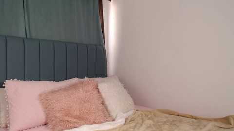 Media: Video of a minimalist bedroom corner with a grey padded headboard, a beige quilted blanket, and a fluffy pink pillow on a bed against a white wall, featuring a partially visible window with a teal curtain.